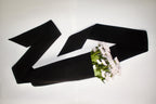 A black flower tote arranged in a loose loop on a plain white background, with a small bouquet of white flowers tucked into one end.