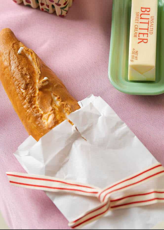 Candle shaped like a loaf of bread wrapped in paper with a red bow on a pink tablecloth.