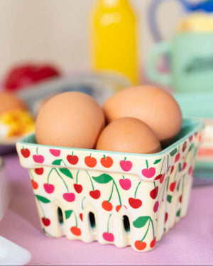 Yellow ceramic basket with pink and red cherries on it holding eggs on a table.