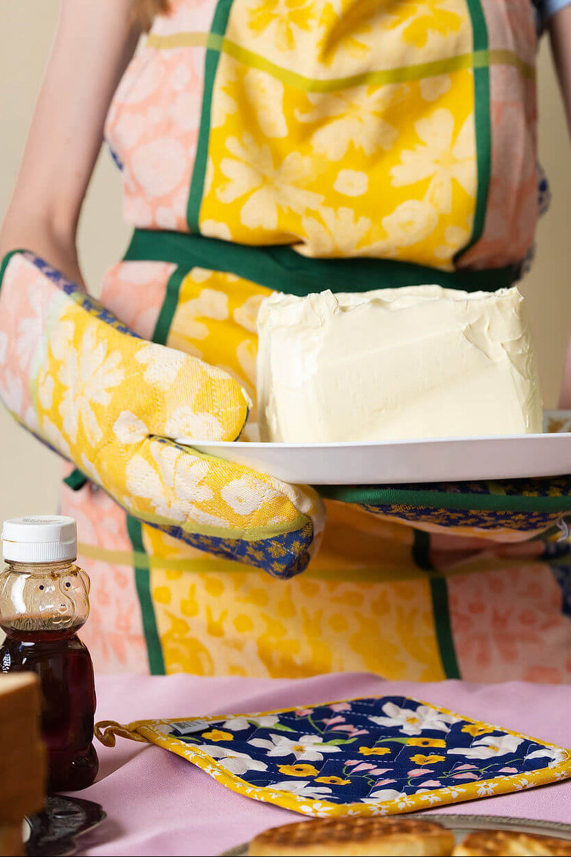 Person wearing a pink, yellow, and green floral apron with a matching hot pad set holding a plate of butter.