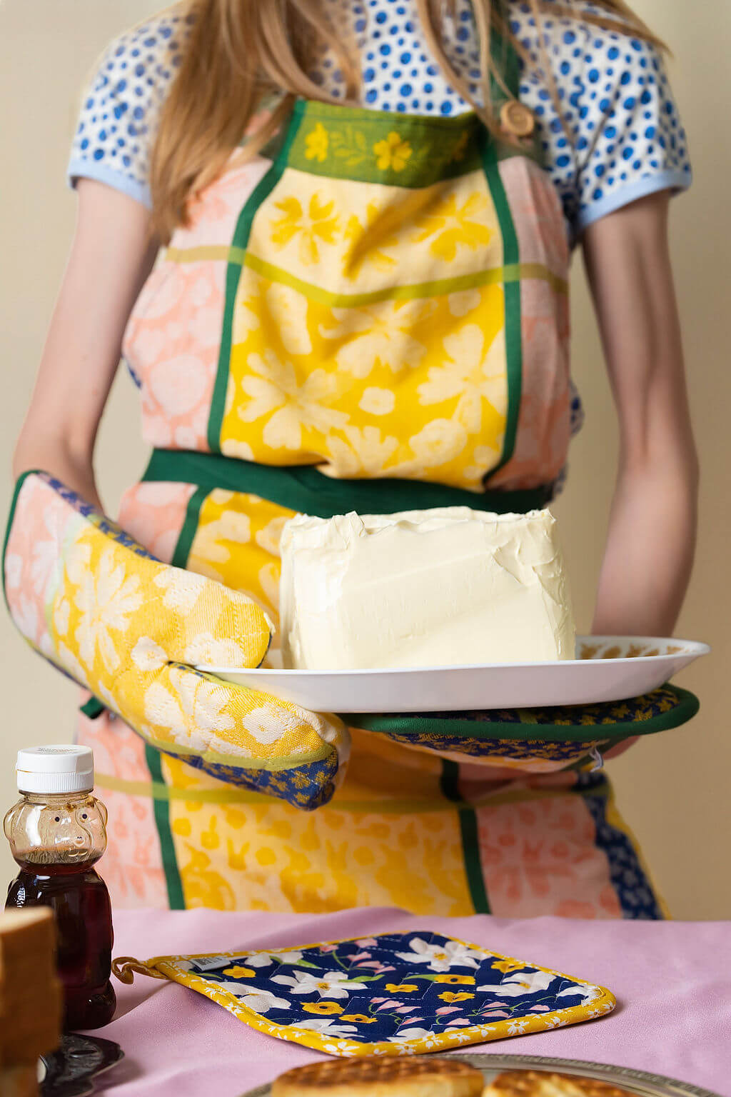Person wearing a yellow, pink and green floral apron and oven mitt, holding a plate of butter.