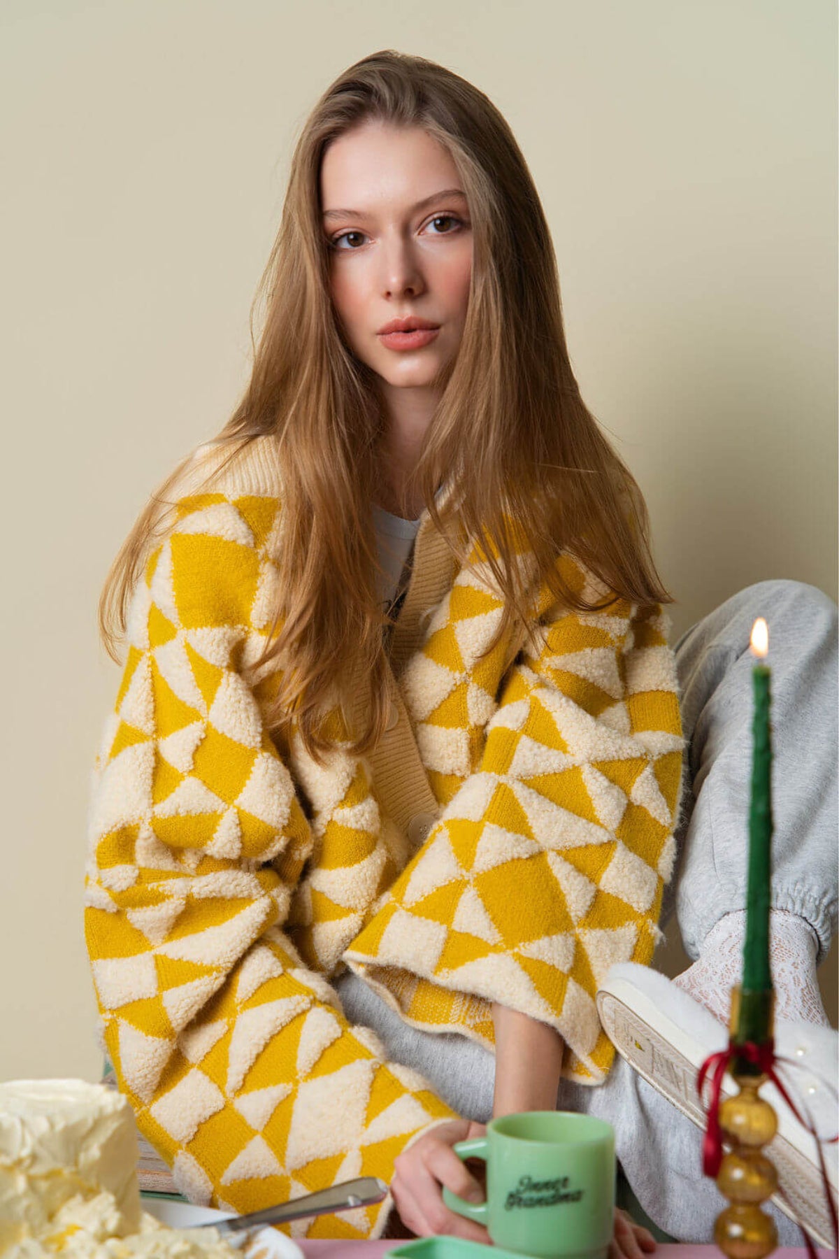 A person wearing a yellow and white patterned quilted cardigan sweater blanket at a table with a candle and green mug.