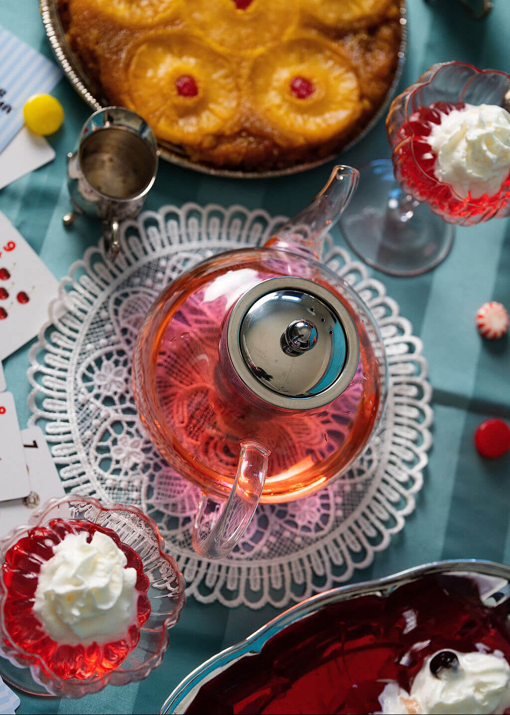 Glass tea pot with stainless steel filter on a doily with desserts at a table.