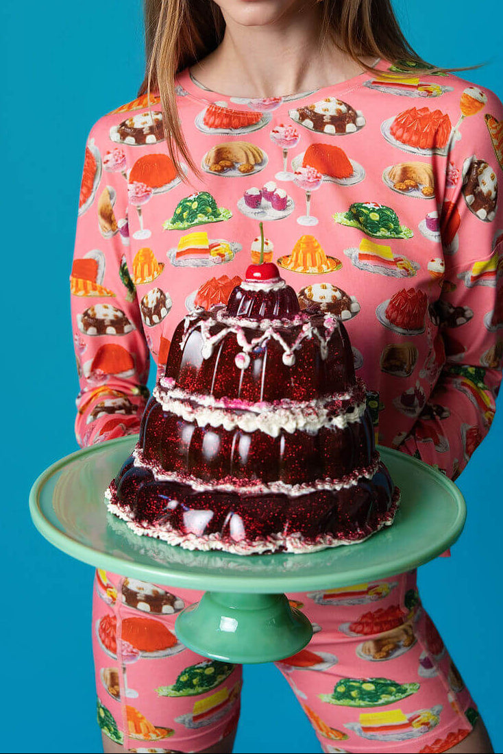 Person wearing a pink set with jello print holding a green jadeite cake stand with a red jello cake dessert.