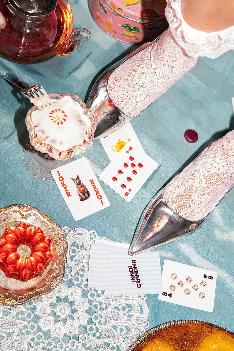 Person wearing lace socks and shoes over a table with tea set and playing cards.