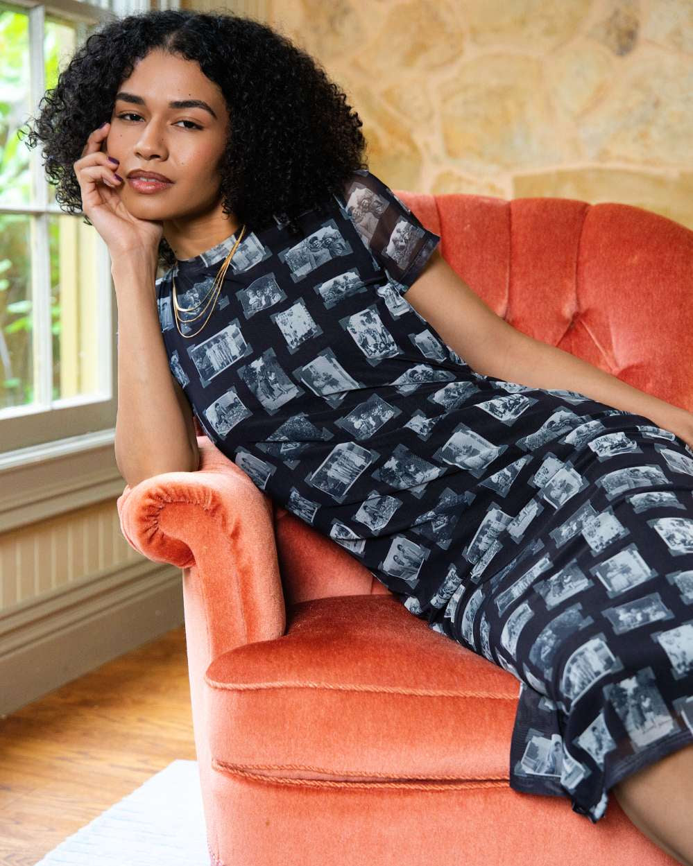 Person wearing black and gray mesh fabric dress with printed black and white images of grandmas, people and objects, siting on a orange couch indoors. 
