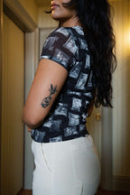 Side of person wearing black and gray mesh printed fabric top of black and white images of grandmas, people and objects with white pants and a tattoo on their arm, standing indoors.