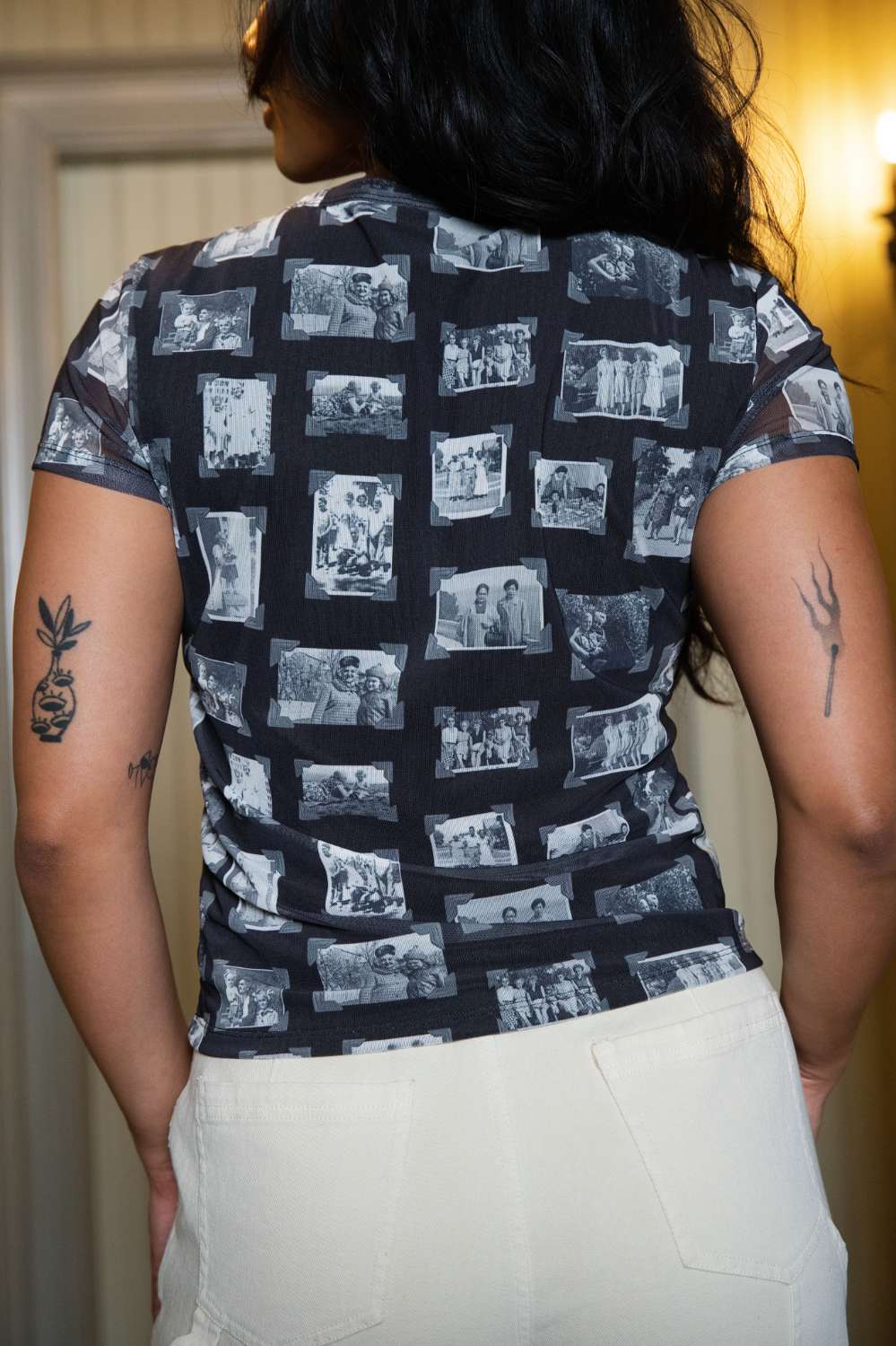 Back of person wearing black and gray mesh printed fabric top of black and white images of grandmas, people and objects with white pants and a tattoo on their arm, standing indoors.