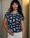 Person wearing black and gray mesh printed fabric top of black and white images of grandmas, people and objects and white pants, standing indoors.