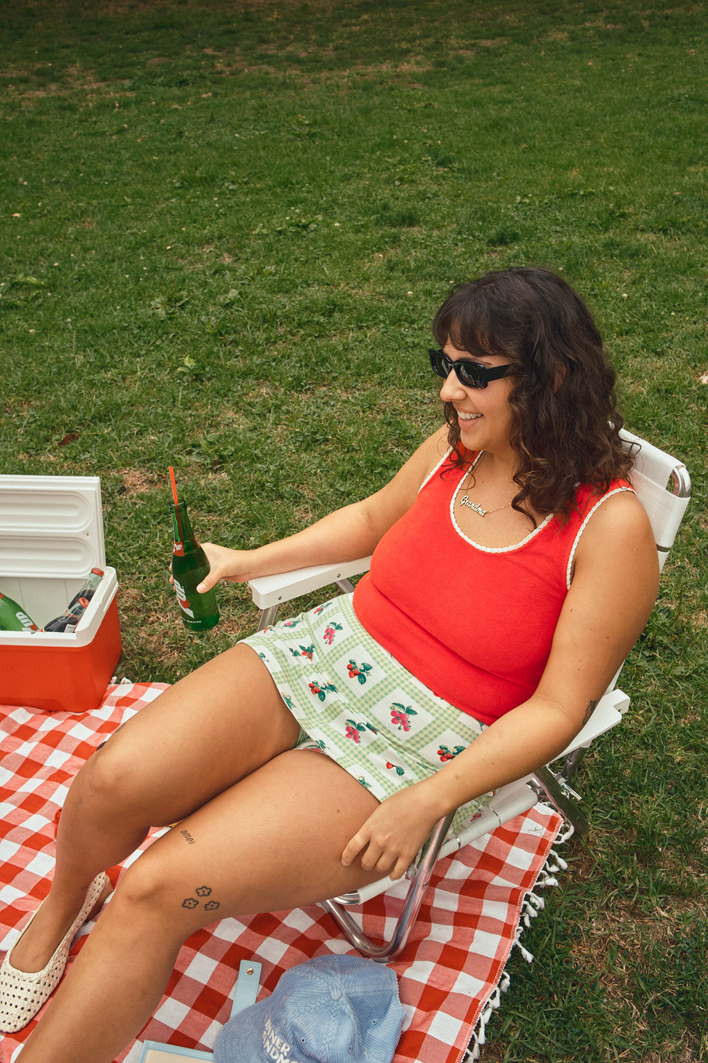 Person wearing a red tank top and green-and-white gingham skort with a fruit print sitting on a lawn chair and picnic blanket.