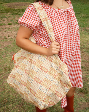 Person wearing a red and white dress holding a tote bag with patterned fabric of vegetable labels of pickles, peppers, tomatoes, asparagus and onions.