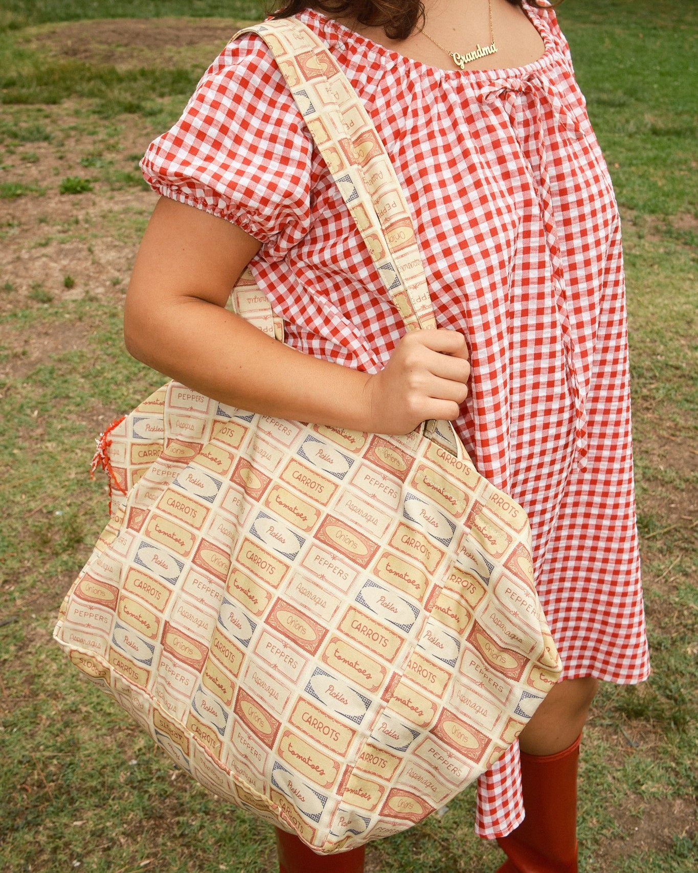 Person wearing a red and white dress holding a tote bag with patterned fabric of vegetable labels of pickles, peppers, tomatoes, asparagus and onions.