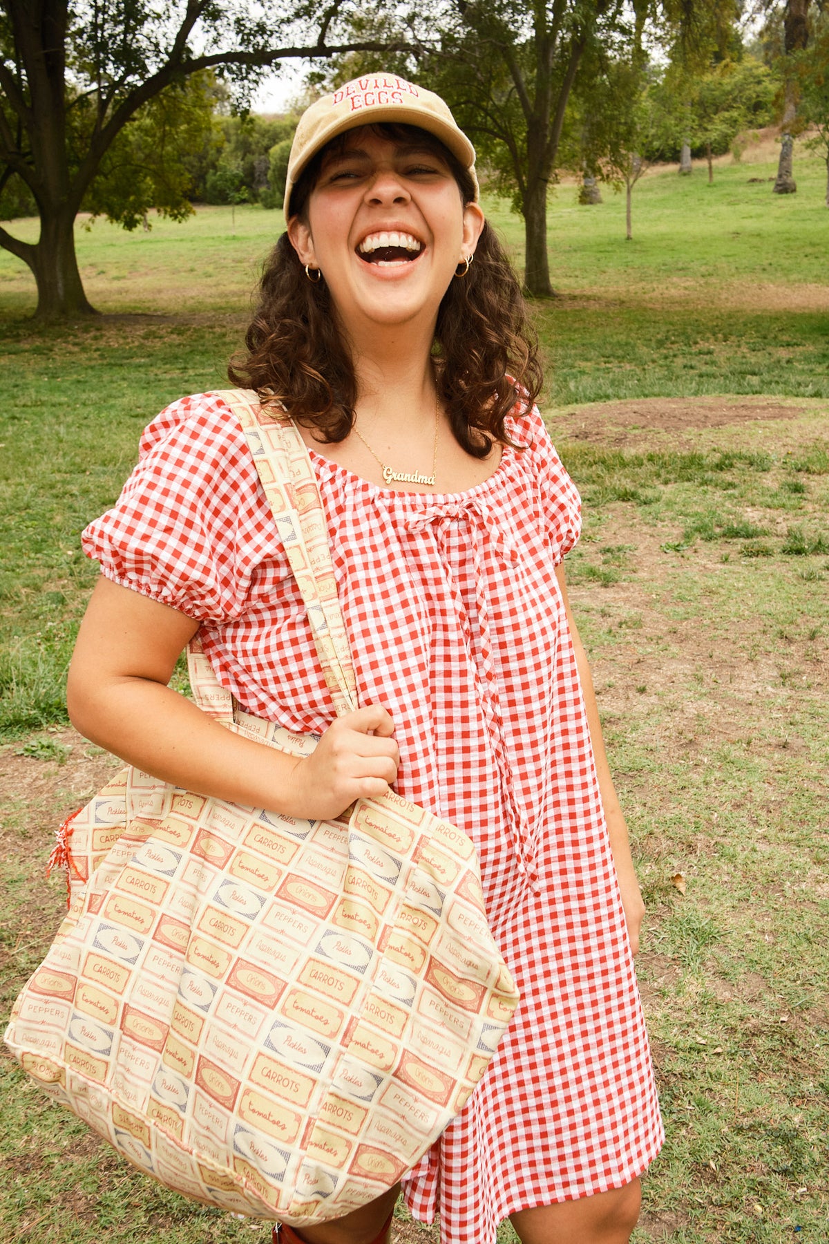 Person wearing a yellow hat and a red-and-white dress, holding a tote bag with a pattern of vintage-style vegetable labels featuring pickles, peppers, tomatoes, asparagus, and onions.