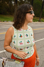Person wearing a printed green and white gingham tank top with fruit on it, wearing sunglasses standing on a road. 