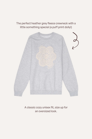 A light heather grey crewneck sweatshirt with a cream puff-print dolly graphic on the chest, shown on a light beige background with descriptive text and a brown arrow.