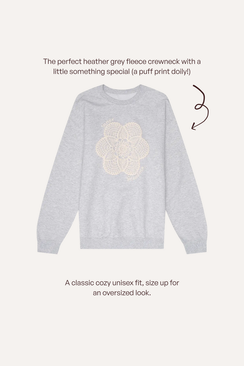 A light heather grey crewneck sweatshirt with a cream puff-print dolly graphic on the chest, shown on a light beige background with descriptive text and a brown arrow.
