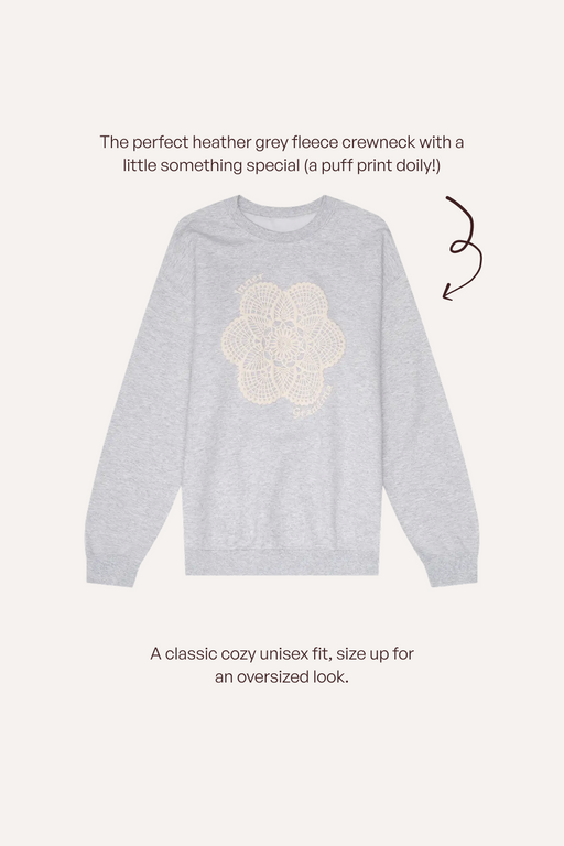 A light heather grey crewneck sweatshirt with a cream puff-print dolly graphic on the chest, shown on a light beige background with descriptive text and a brown arrow.