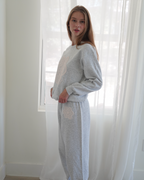 Person wearing a light gray heathered lounge set consisting of a sweatshirt and matching sweatpants with white doily details, standing in a bright room with white sheer curtains 