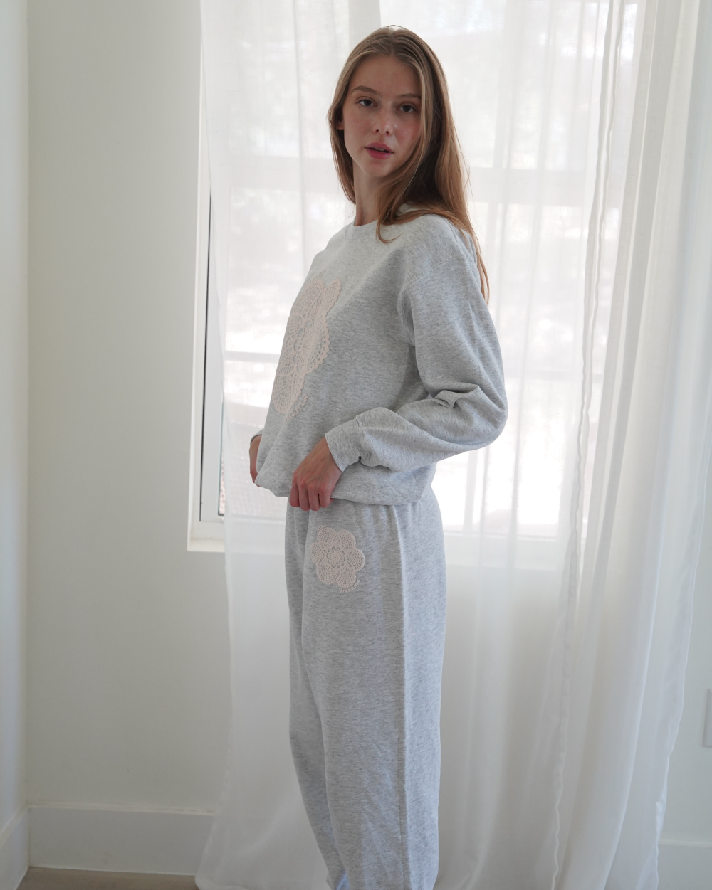 Person wearing a light gray heathered lounge set consisting of a sweatshirt and matching sweatpants with white doily details, standing in a bright room with white sheer curtains 