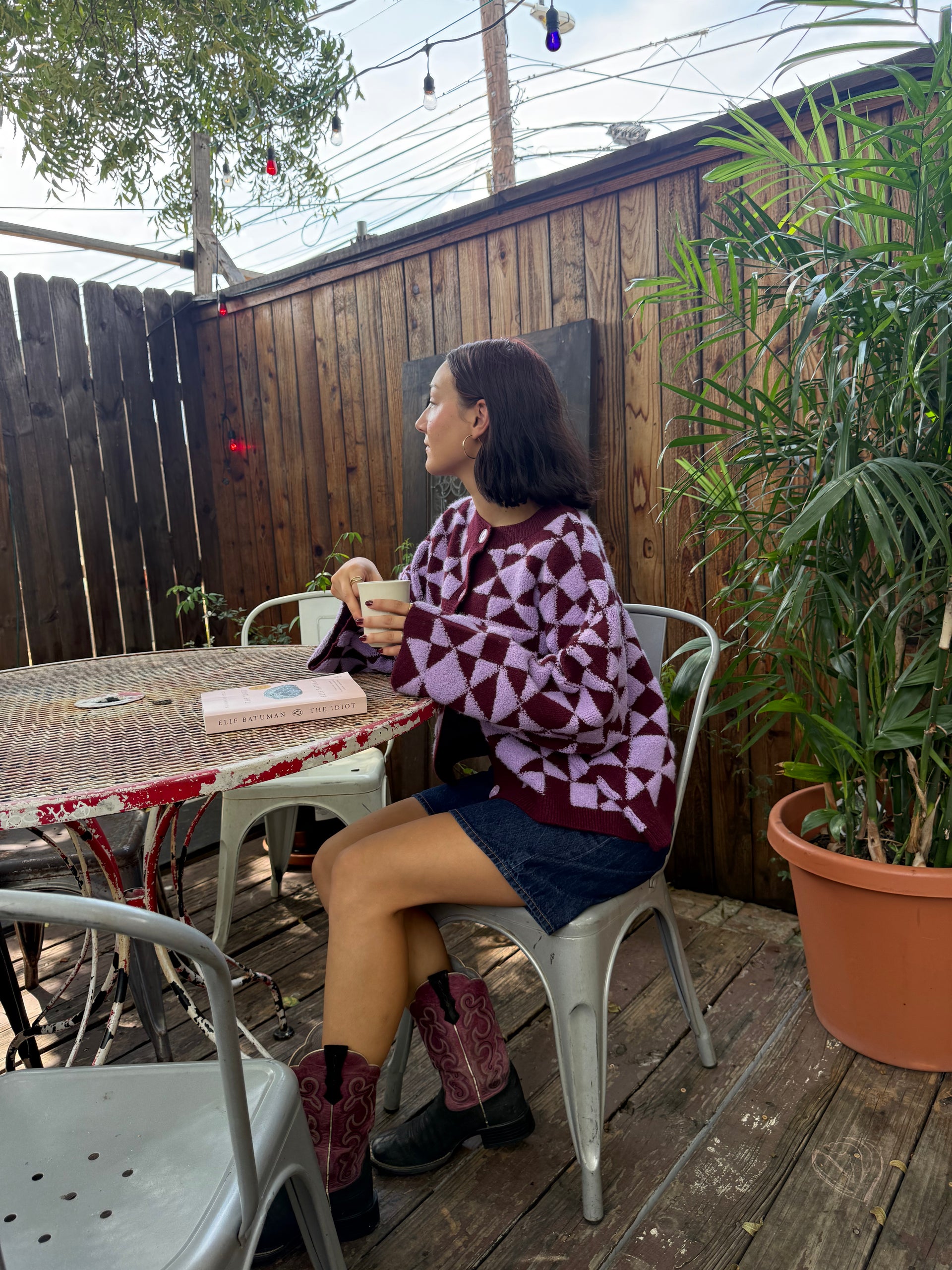 Person sitting drinking coffee wearing a plum and light purple geometric patterned cardigan paired with jean shorts, and purple and black cowboy boots.