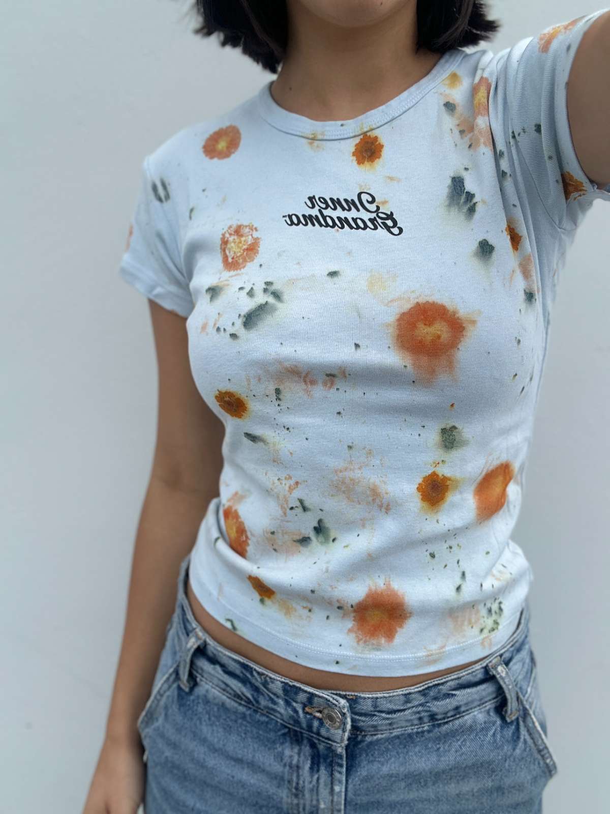 Person wearing a light blue cropped t-shirt with orange and yellow pattern naturally dyed by flowers with "Inner Grandma" text