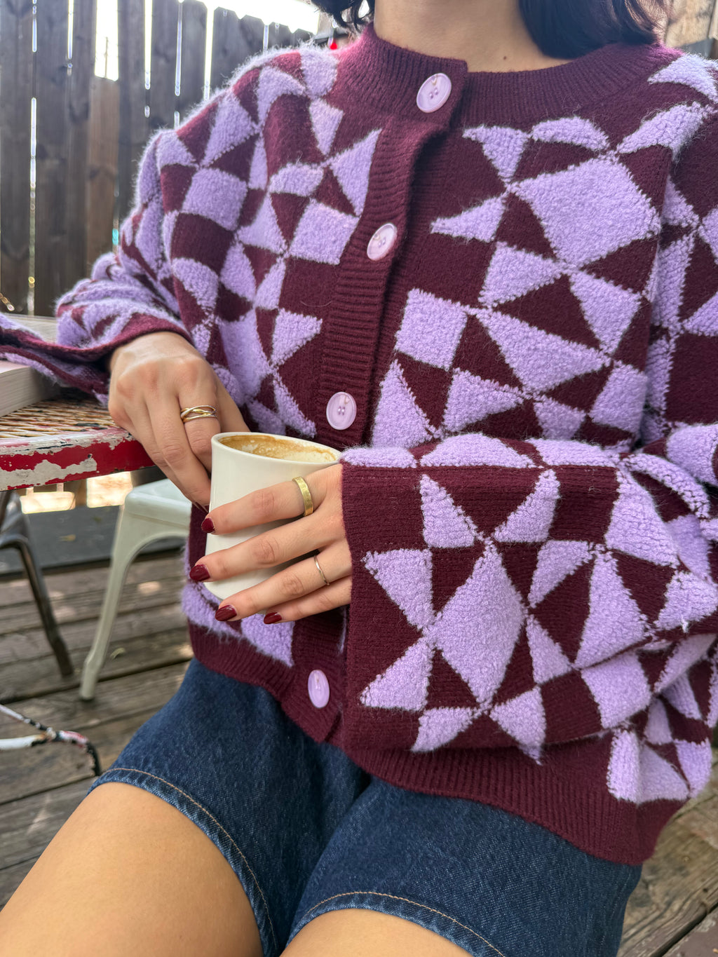 Close up of person wearing a plum and light purple geometric patterned cardigan with purple buttons, sitting drinking coffee.