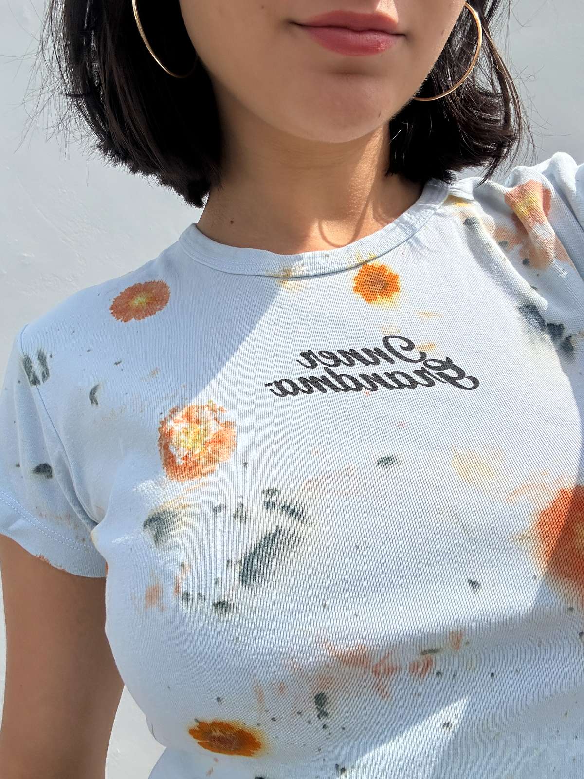 Person wearing a light blue cropped t-shirt with orange and yellow pattern naturally dyed by flowers with "Inner Grandma" text