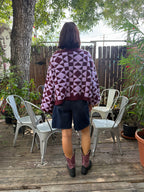 Back of person modeling a plum and light purple geometric patterned cardigan, paired with jean shorts and purple and black cowboy boots.