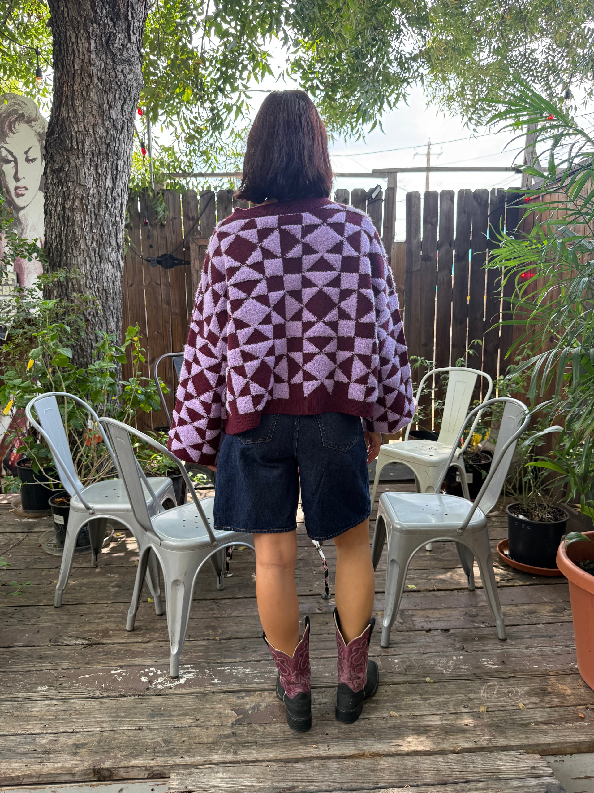 Back of person modeling a plum and light purple geometric patterned cardigan, paired with jean shorts and purple and black cowboy boots.