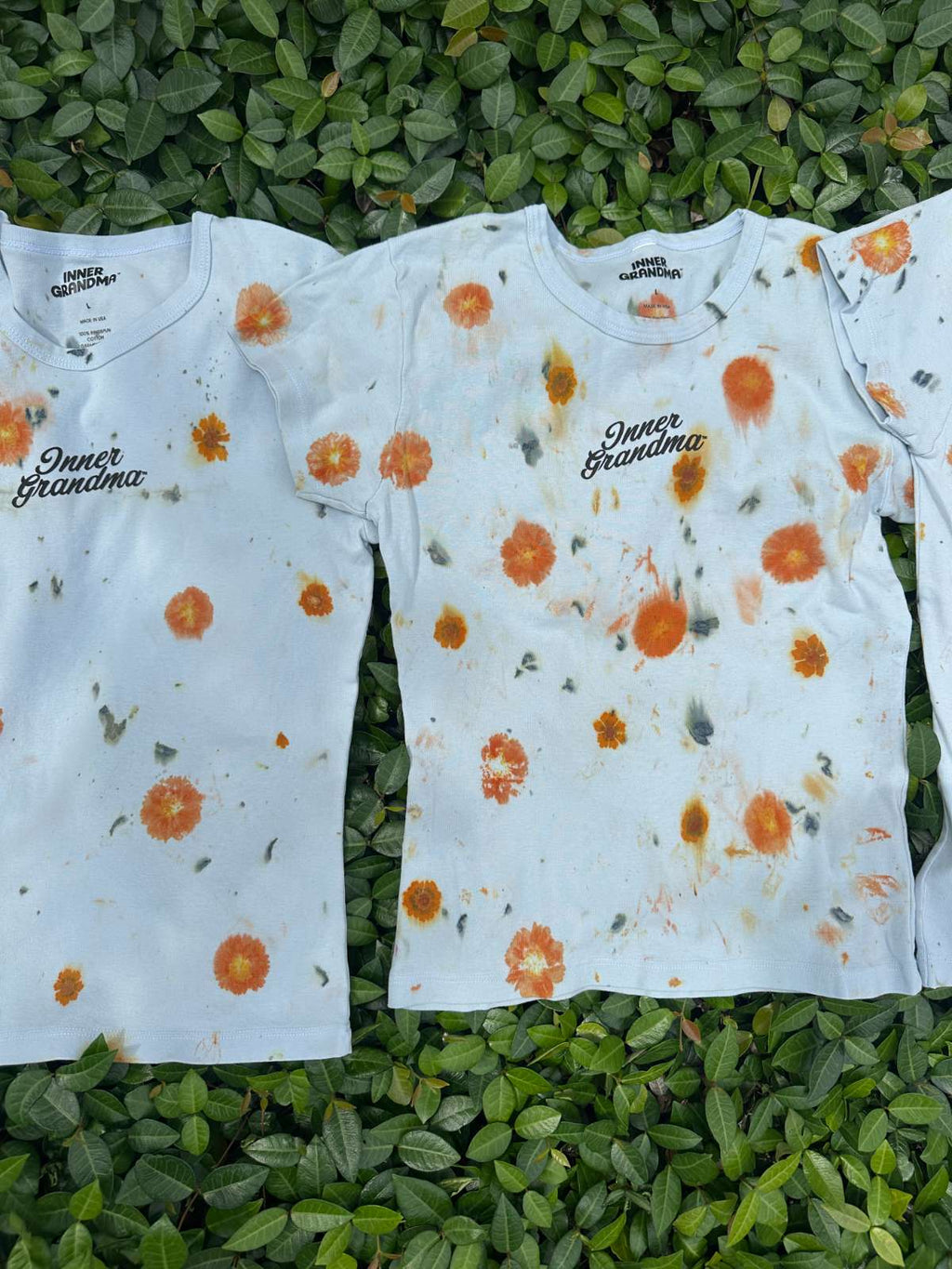 Light blue cropped t-shirts with orange and yellow pattern naturally dyed by flowers with "Inner Grandma" text, blue jeans and green plants in the background on green bush