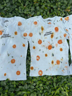 Light blue cropped t-shirts with orange and yellow pattern naturally dyed by flowers with "Inner Grandma" text, blue jeans and green plants in the background on green bush