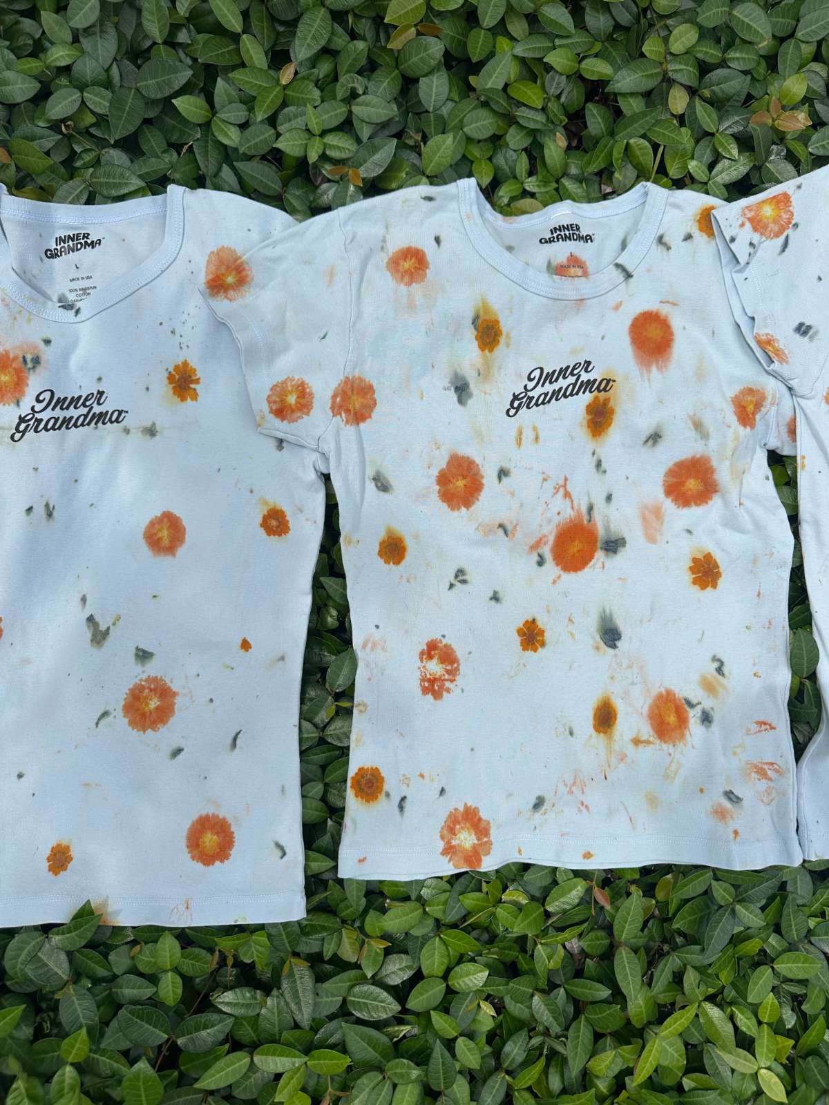 Light blue cropped t-shirts with orange and yellow pattern naturally dyed by flowers with "Inner Grandma" text, blue jeans and green plants in the background on green bush