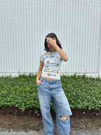 Person wearing a light blue cropped t-shirt with orange and yellow pattern naturally dyed by flowers with "Inner Grandma" text with jeans and green plants