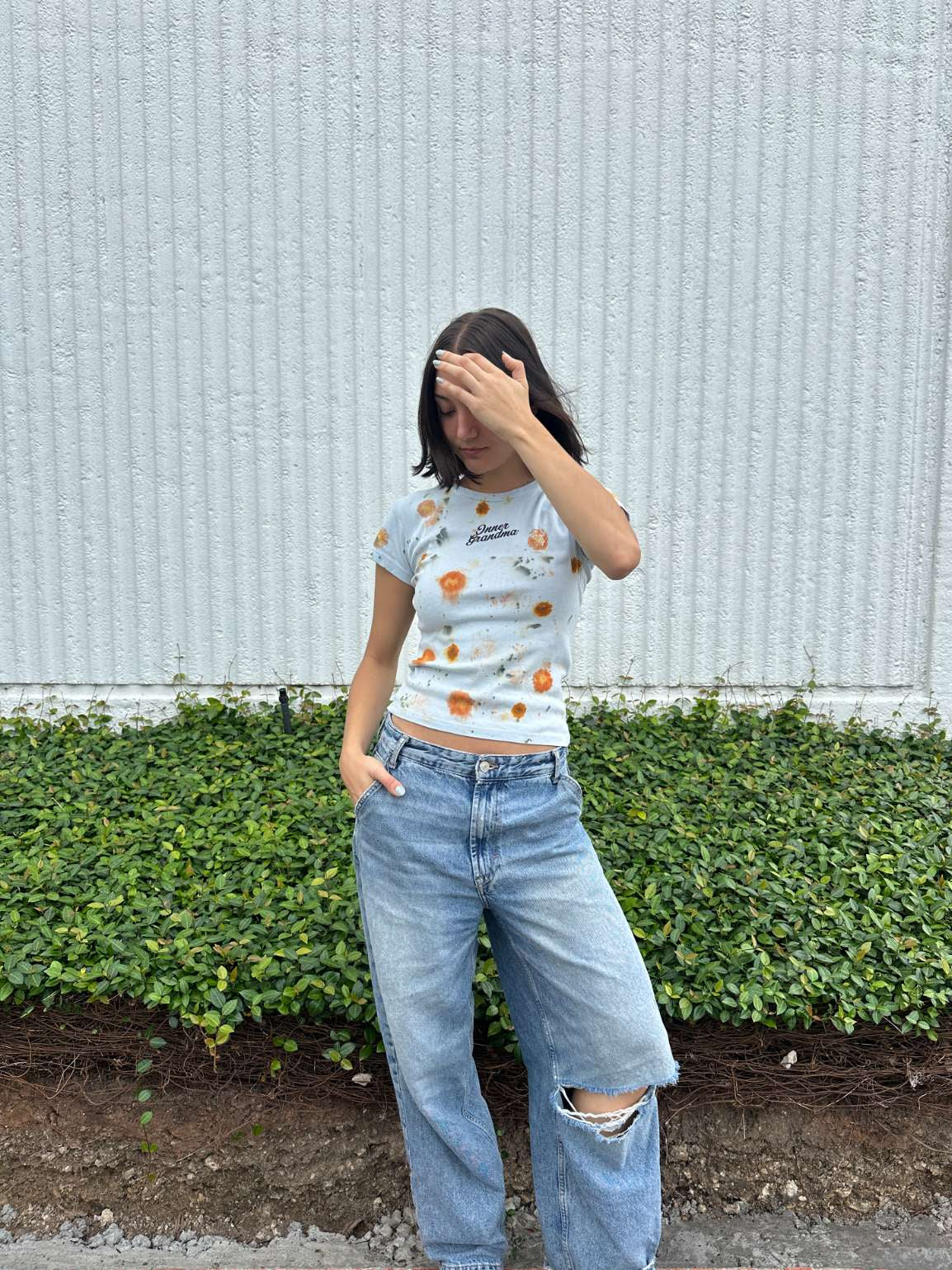 Person wearing a light blue cropped t-shirt with orange and yellow pattern naturally dyed by flowers with "Inner Grandma" text with jeans and green plants