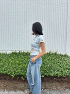 Person wearing a light blue cropped t-shirt with orange and yellow pattern naturaly dyed by flowers with "Inner Grandma" text, blue jeans and green plants in the background