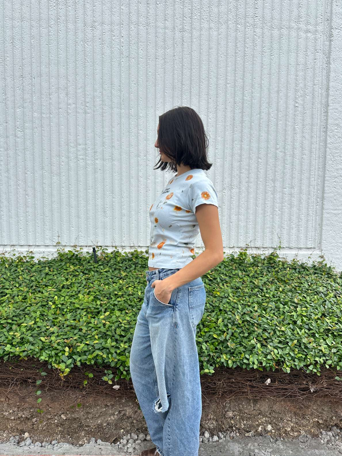 Person wearing a light blue cropped t-shirt with orange and yellow pattern naturaly dyed by flowers with "Inner Grandma" text, blue jeans and green plants in the background