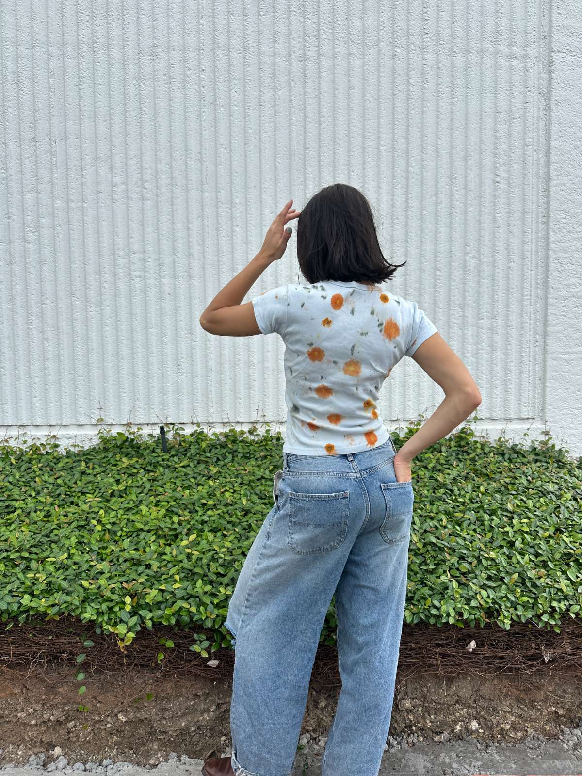 Person wearing a light blue cropped t-shirt with orange and yellow pattern naturaly dyed by flowers with "Inner Grandma" text, blue jeans and green plants in the background