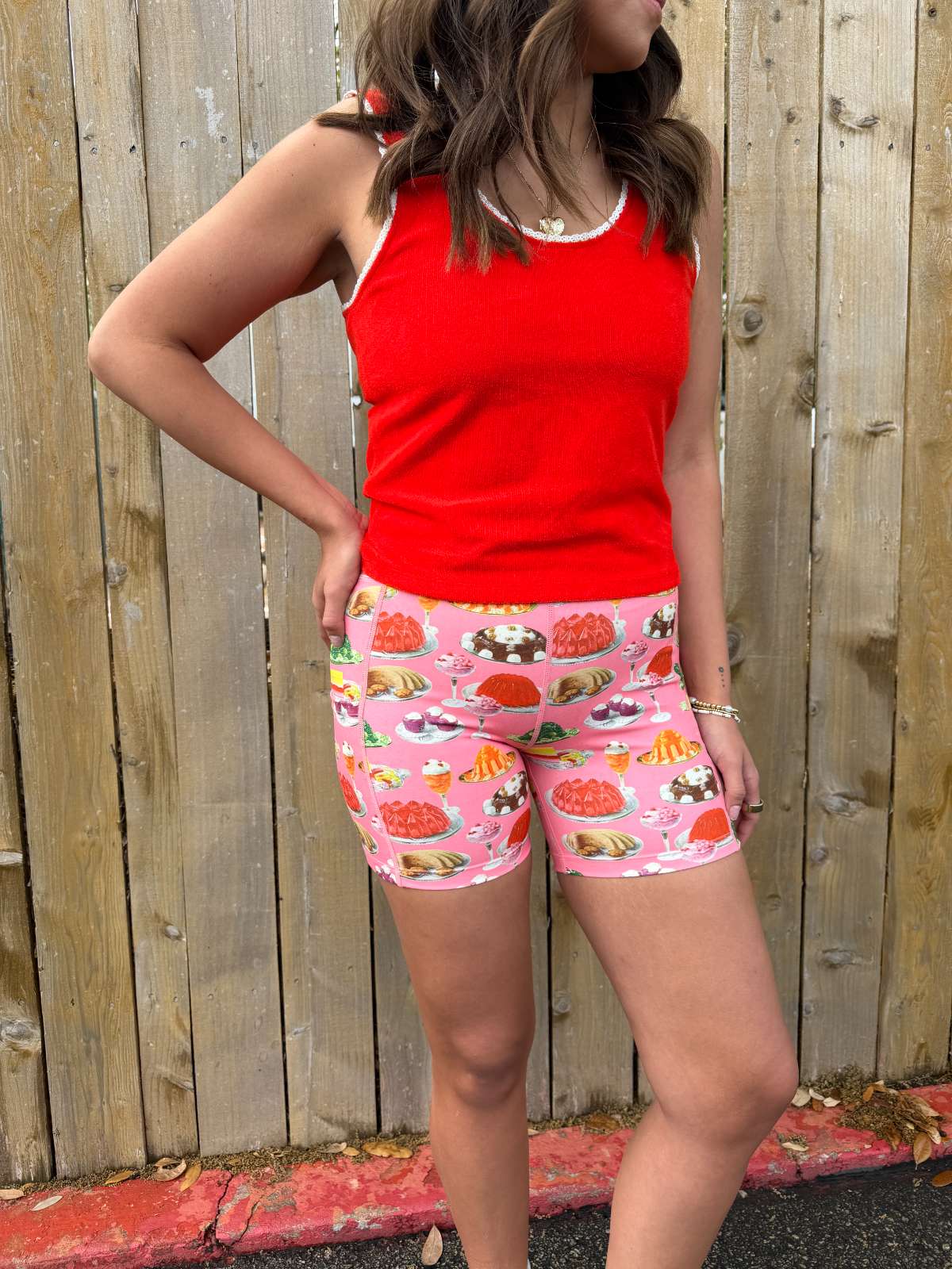 Person wearing a red tank top and pink shorts with dessert print against a wooden fence.
