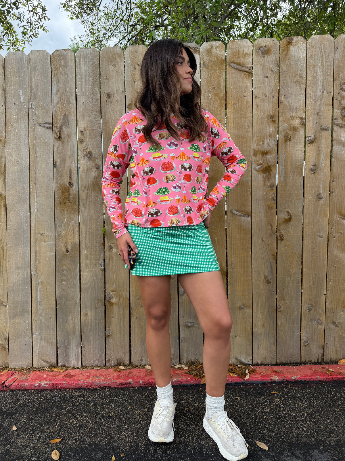 Person wearing a pink long sleeve sun shirt with a dessert print and a green gingham dress standing against a wooden fence with a natural background outside