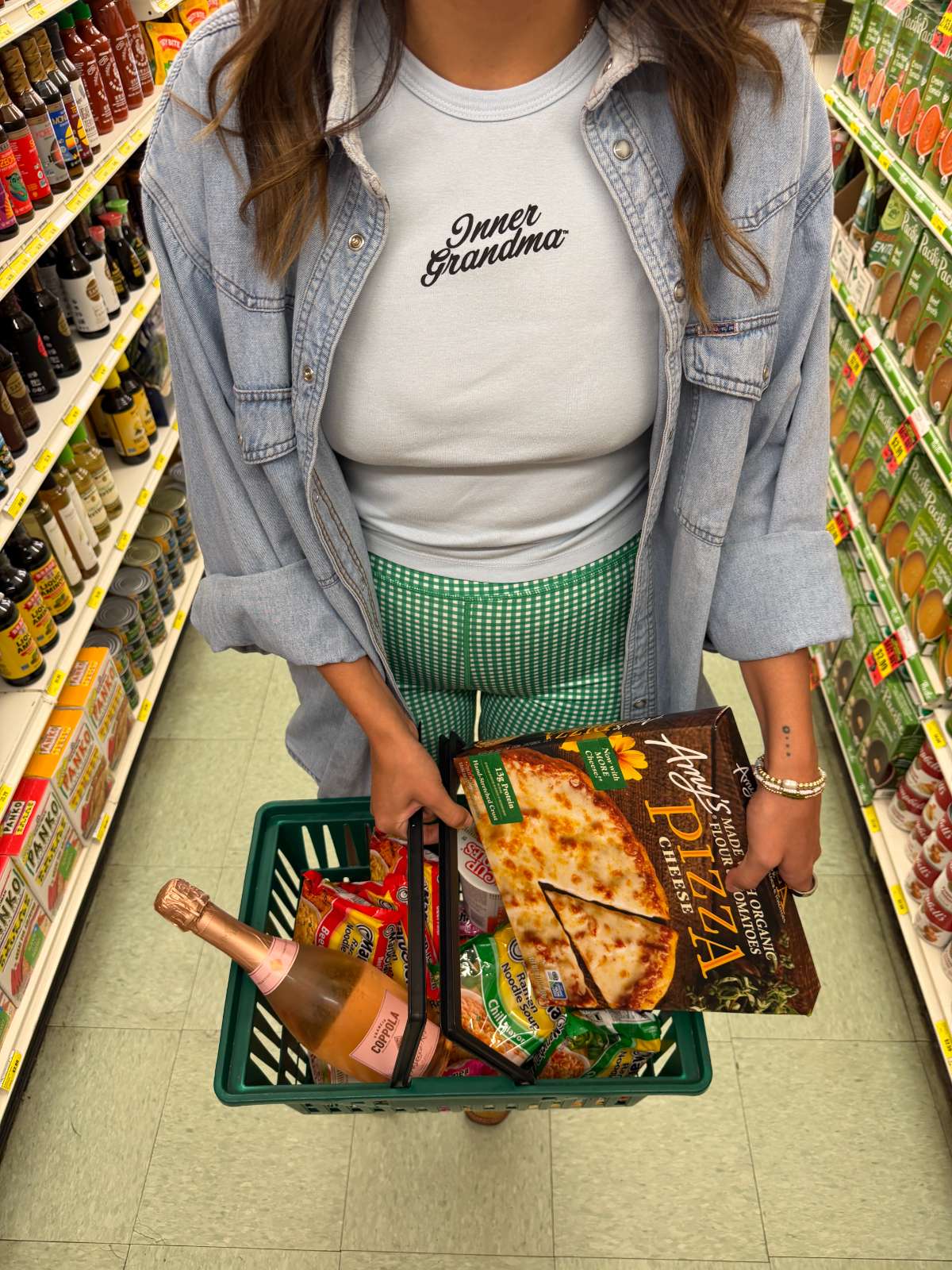 Person shopping in a supermarket aisle with a basket full of items in a light blue Inner Grandma cropped t-shirt and green gingham shorts.