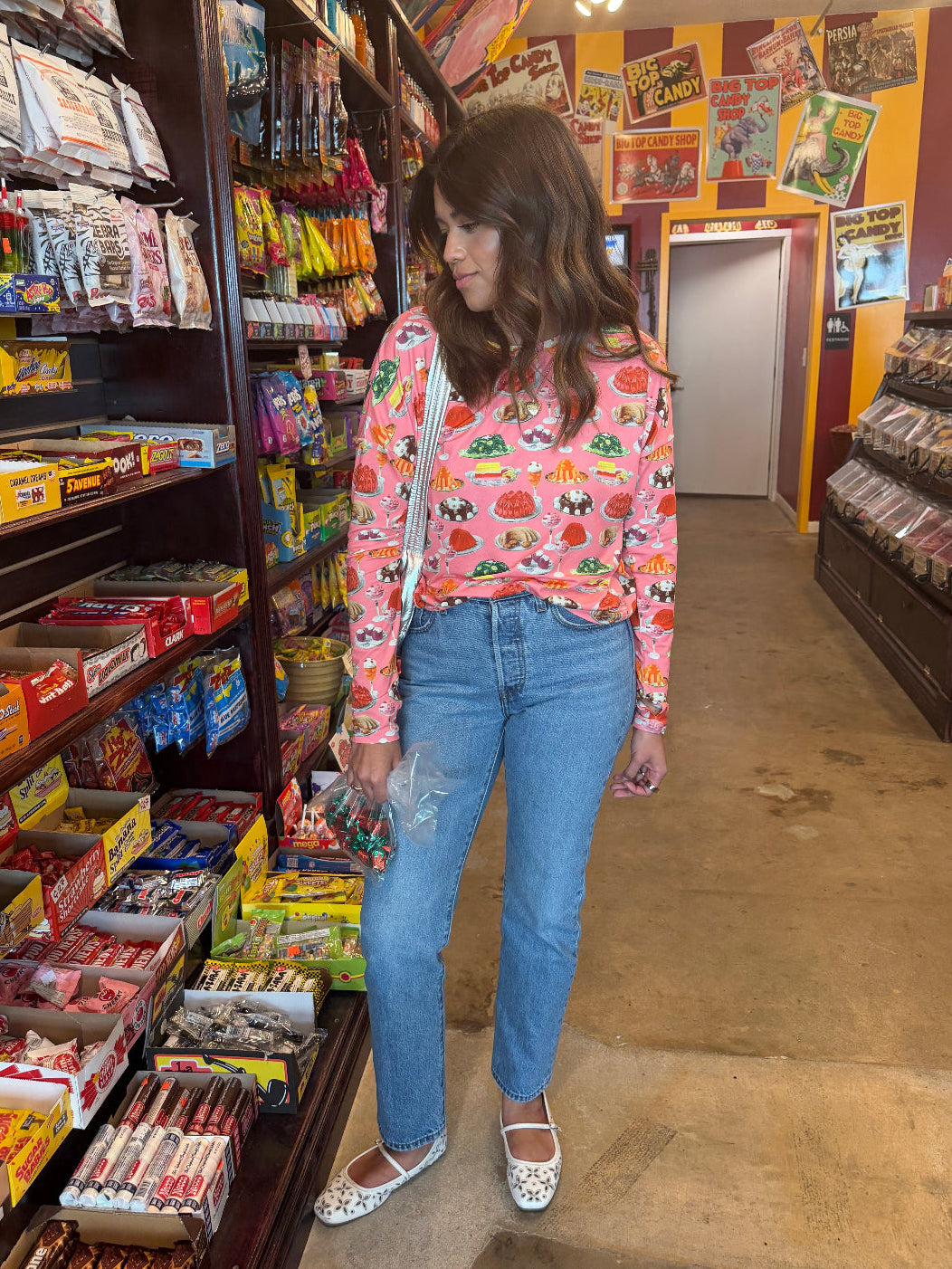 Person wearing a pink long sleeve sun shirt with a dessert print and jeans standing in a candy store with shelves stocked with various items.