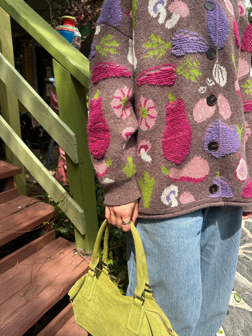 A close-up of a person wearing a chunky knit cardigan sweater with a multicolor vegetable pattern on a brown base with jeans and a green purse.