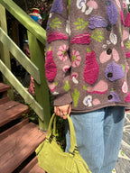 A close-up of a person wearing a chunky knit cardigan sweater with a multicolor vegetable pattern on a brown base with jeans and a green purse.