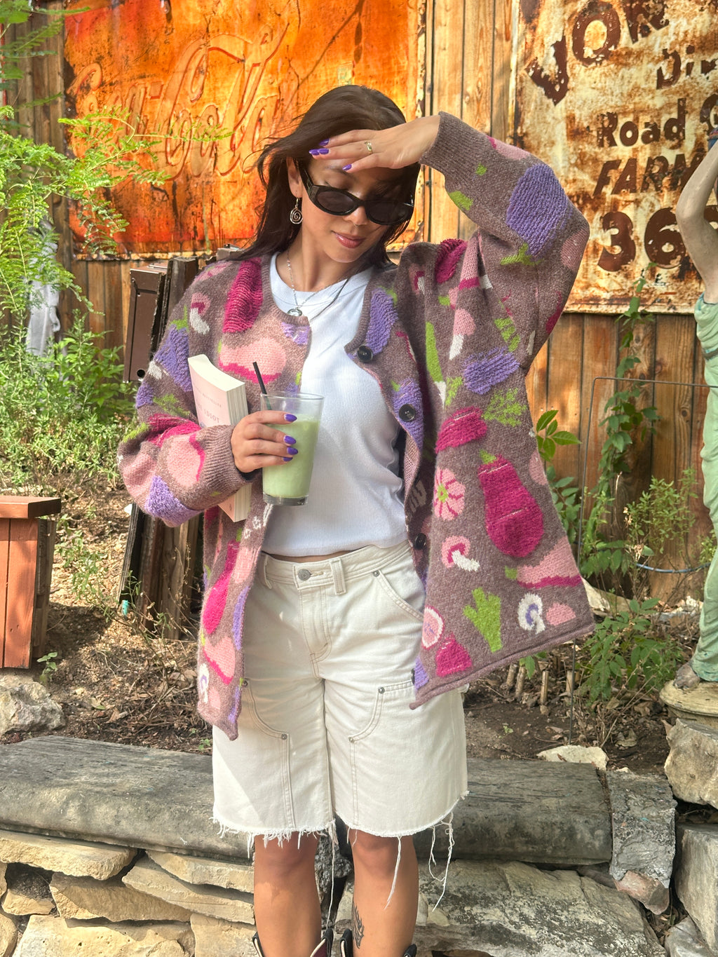 Person wearing a chunky knit cardigan sweater with a multicolor vegetable pattern on a brown base with a white shirt and while shorts, holding a matcha outdoors.