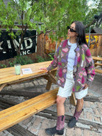 A person posing outdoors at a wooden picnic table, wearing a colorful patterned sherpa zip-up jacket, a white t-shirt, white denim shorts, and purple and black cowboy boots.