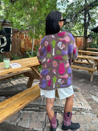 A person standing outdoors at a wooden picnic table, wearing a brown knit cardigan with colorful abstract mushroom and floral motifs in pink, purple, green, and white, paired with white shorts and purple cowboy boots.
