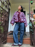 Person stands on a porch wearing a purple and burgundy geometric pattern knit cardigan, blue wide-leg jeans, and burgundy shoes.