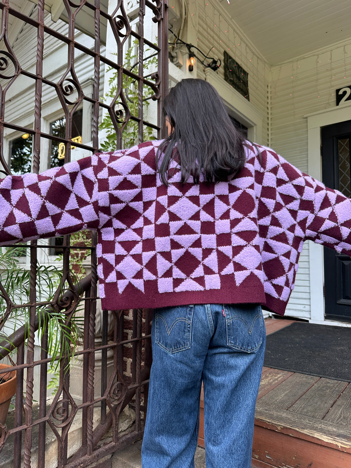 A person viewed from behind wearing a purple and burgundy geometric patterned knit sweater and blue jeans, standing on a porch in front of a decorative metal gate.