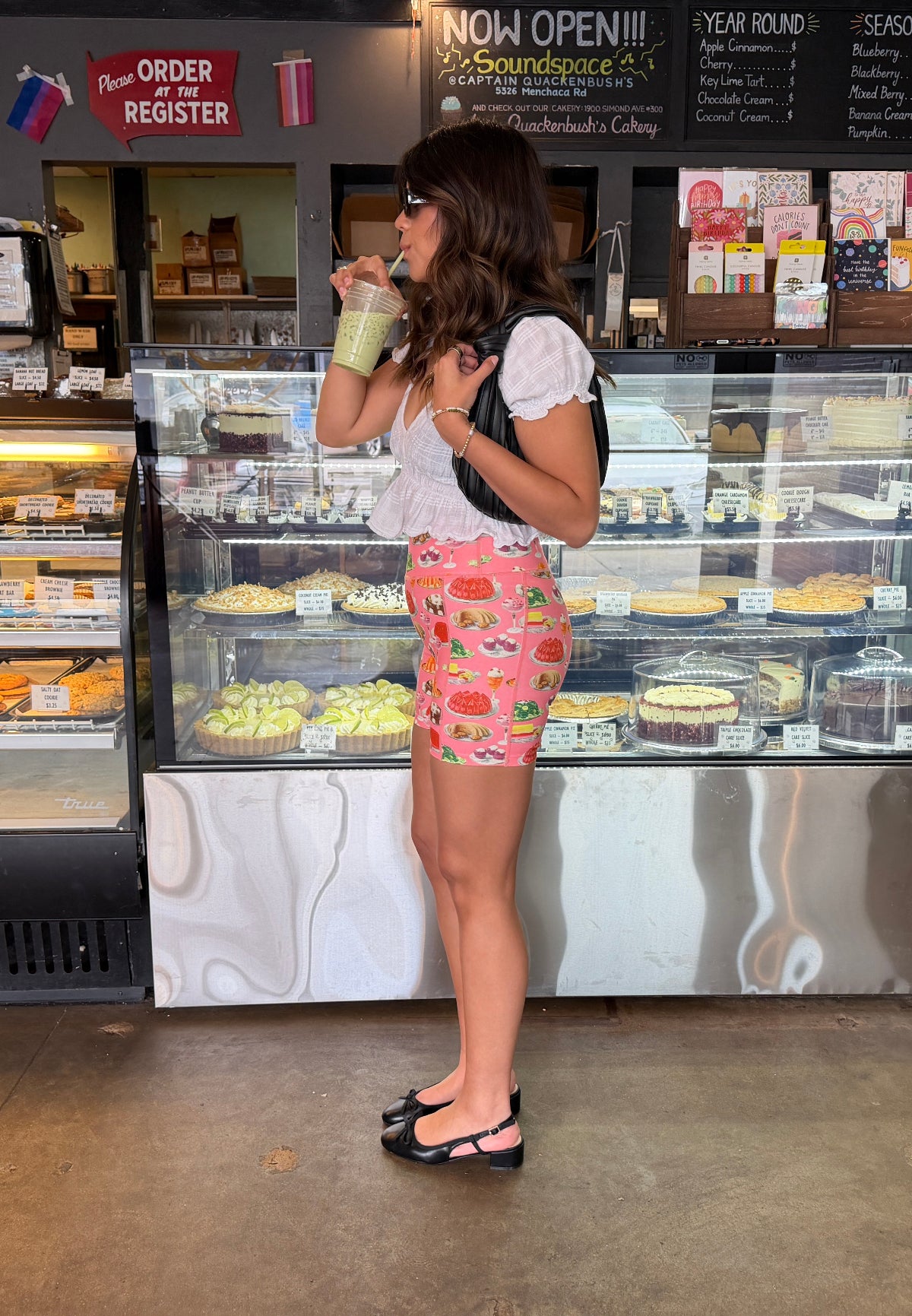 Person wearing a white shirt and pink casual shorts with a dessert print standing in front of a bakery display case at a coffee shop, holding a drink.