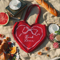 Red heart-shaped bag with 'Book Lover' text on a blanket with food and books.
